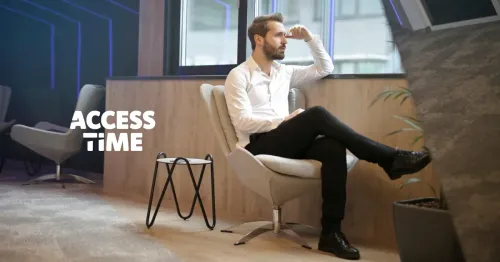 A man in a white shirt and black pants, sitting in a chair by the window, staring in the window thinking.